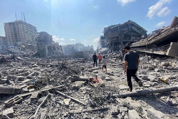 Image of Palestinians walking through destruction in Gaza City: the foreground is completely rubble, while far in the background you can see more buildings that are either collapsed or badly damaged. The sky is blue and clear. 