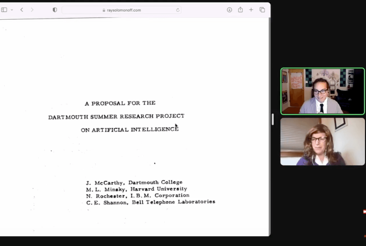 Screenshot of a Zoom meeting with two participants. On the right is a screenshared document that looks like it was written on a typewriter. It reads in all caps, "A proposal for the Dartmouth Summer Research Project on artificial intelligence. J. McCarthy, Dartmouth College. M.L. Minsky, Harvard University. N. Rochester, I.B.M. Corporation. C.E. Shannon, Bell Telephone Laboratories." On the right are the thumbnails of the participants: Alex Hanna, an Arab woman with curly black hair pulled back. She is wearing thick, black-framed glasses, a gray suit jacket, white collared shirt and black tie. The room behind her is covered in art. On the bottom is Emily M. Bender, a white woman who is wearing a long, straight brown wig. She also wears a collared white shirt under a purple cardigan sweater, and glasses. The room behind her has a white wall and closed off-white window shades.