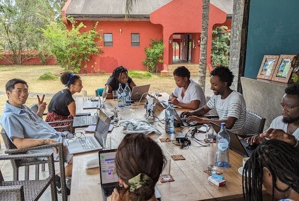 DAIR team members on their laptops around a big wooden table in an outdoor meeting. There is a palm tree in the background, and the walls of the house behind them are red. On the left, Nathan is looking into the camera and signing a peace sign.