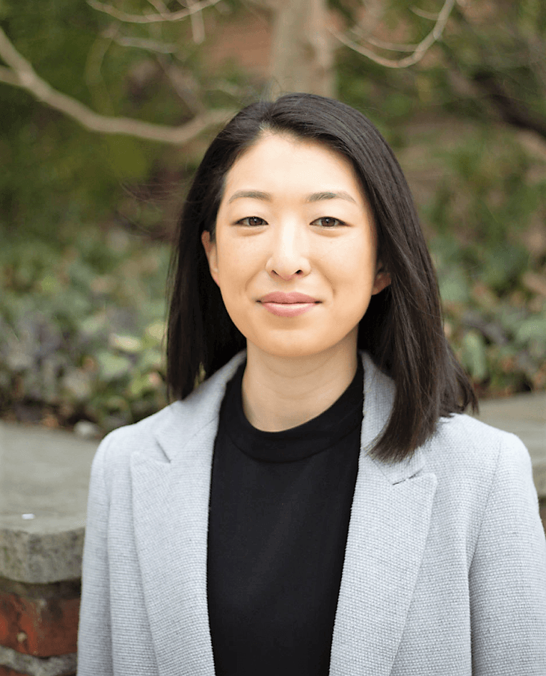 Photo of an Asian woman with straight black hair down to her shoulders. She wears a gray blazer and a blank shirt. She is standing outside, with blurry vegetation and tree branches behind her.