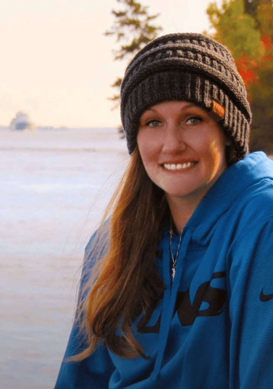 Krystal, a white woman with long brown hair, looking into the camera. She is wearing a bini and a blue hoodie. In the background is what looks like an ocean/sea, and some greenery.