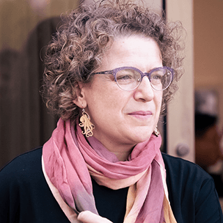 Photo of a white woman from the shoulders up. She has loosely curled brown hair, and is looking to the right edge of the frame. She wears glasses, octopus earrings, and has a pink scarf around her neck. She appears to be standing in front of a wooden door that is partway open into an out of focus room behind her.