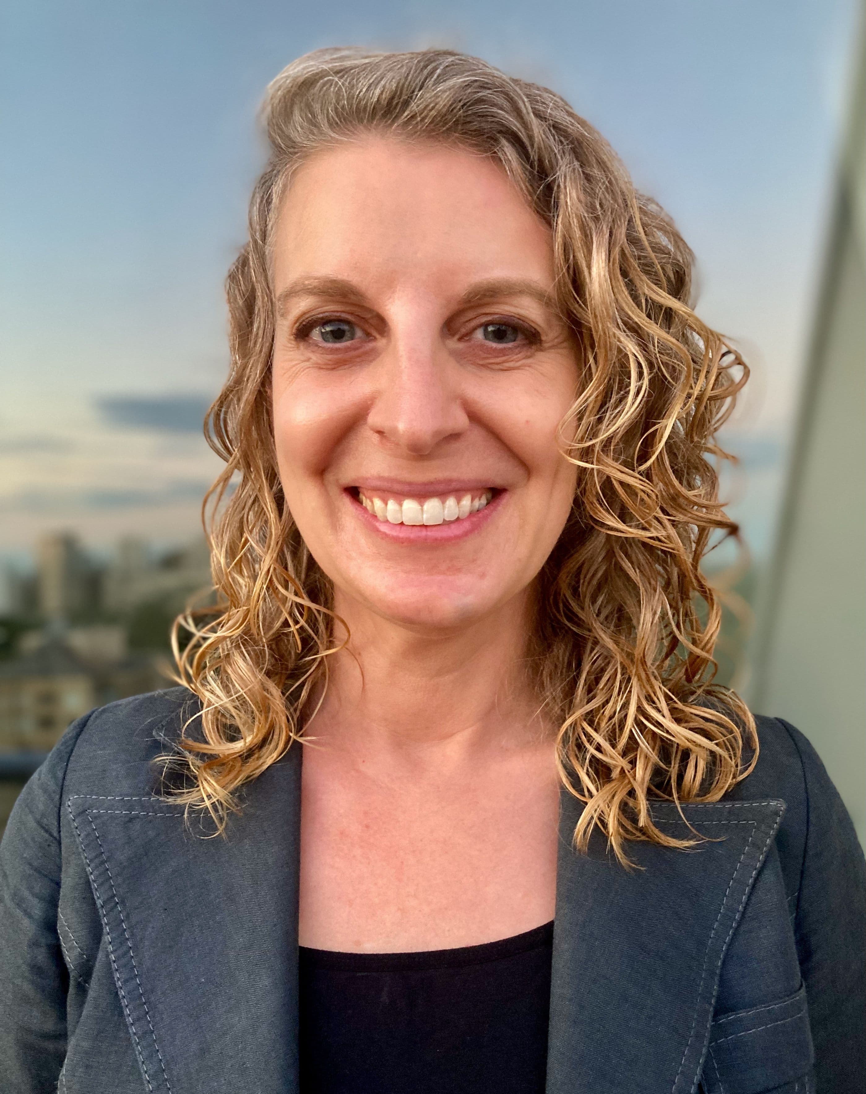 Photo of a white woman from the shoulders up. She has loosely curled shoulder-length blond hair, and is looking at the camera and smiling. She wears a gray blazer and a black shirt under it. She appears to be standing in a high structure, with an out of focus view of a city behind her.