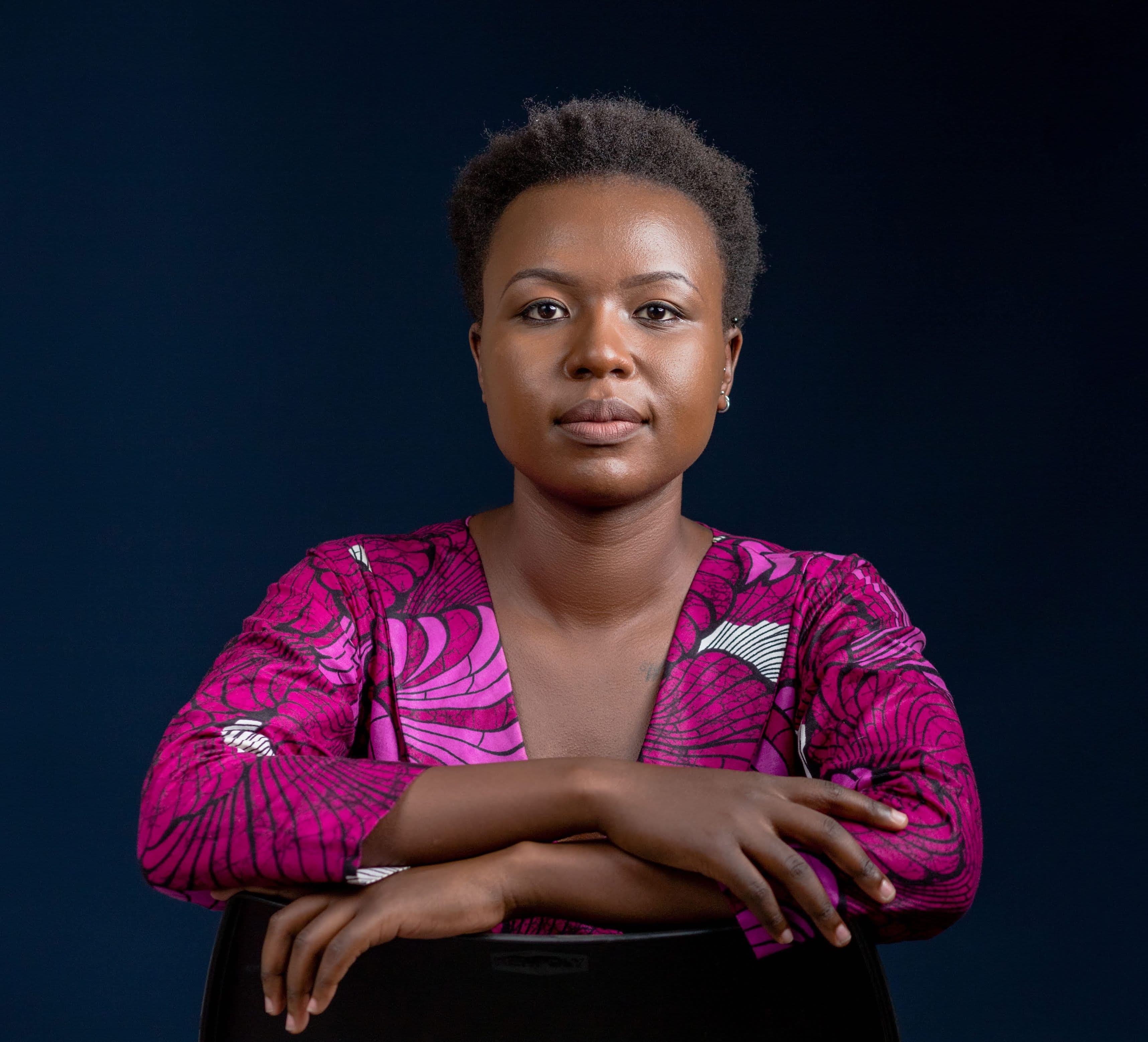 A headshot of Kathleen, a Black woman with short hair, wearing a magenta patterned shirt. She is sitting facing backwards on a chair, with her arms crossed across the back of it like a table. Behind her is a dark blue and black background.