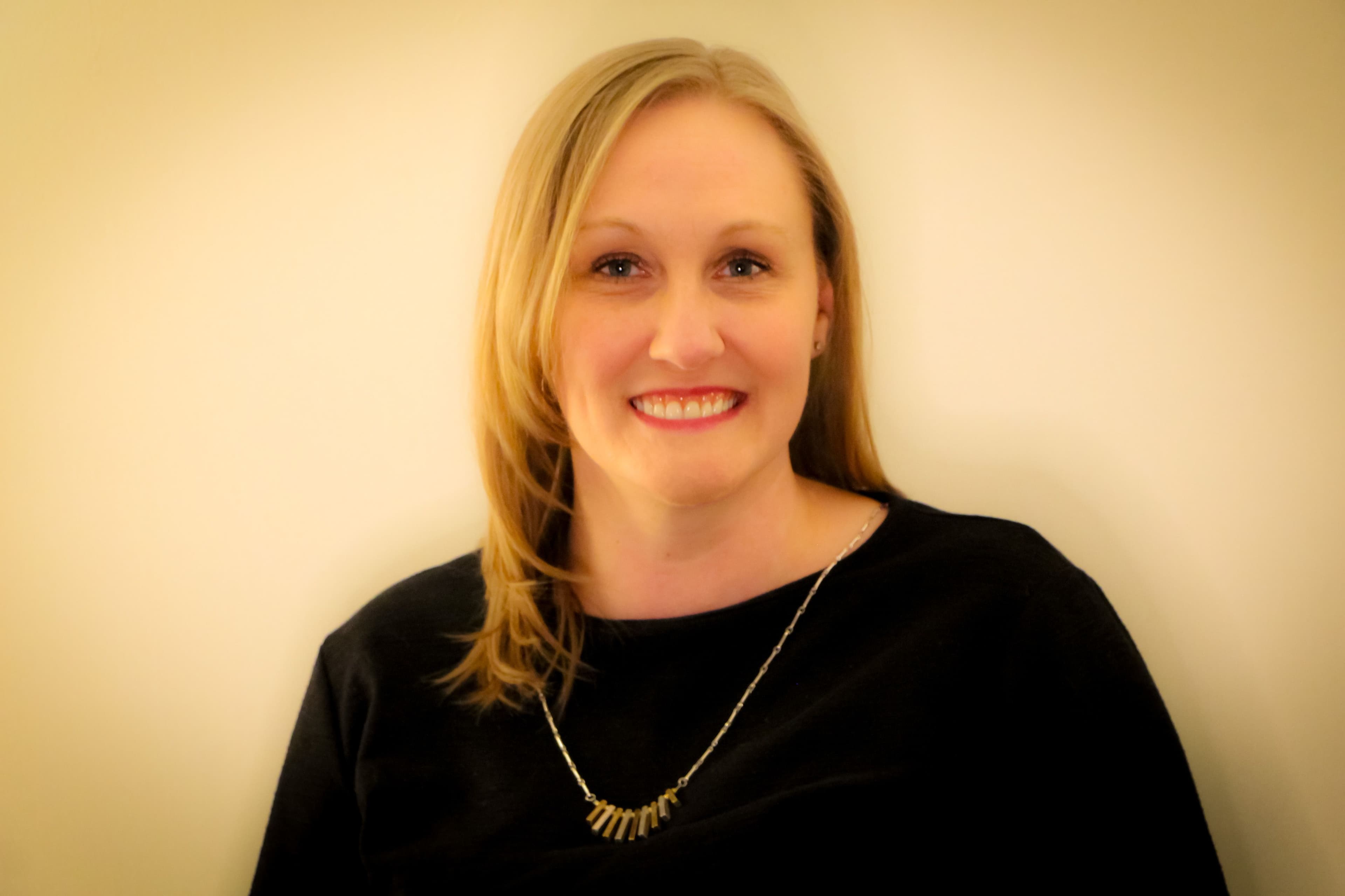 Krystal, a white woman with long brown hair, looking into the camera. She is wearing a black shirt and has a long necklace on. In the background is a neutral off-white wall.