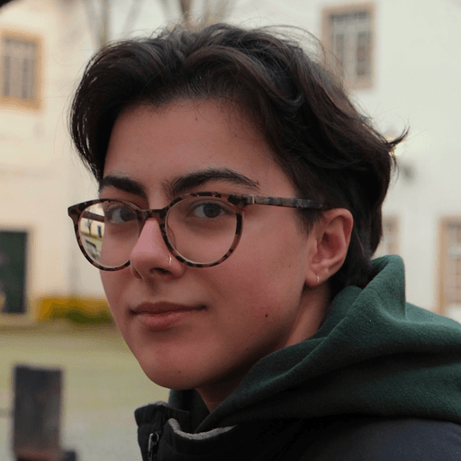 Photo of Francesca from the shoulders up. Francesca is a masculine-presenting person with straight, ear-length brown hair, a small nose ring, and glasses. Francesca is wearing a shirt with a green hood. The photo captures Francesca from the side, with head turned toward the camera. In the background is the exterior of a a building or house with white walls.