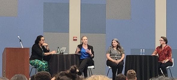 Screenshot of Alex and Krystal sitting on stage at a round table, and two other white woman next to them sitting at another round table.