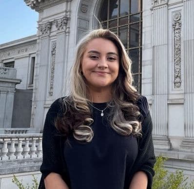 Headshot of Sandra, a Latinx woman with long blond and dark brown hair, wearing a navy blue top with a black cardigan and a silver necklace, with a building with classical architecture in the background