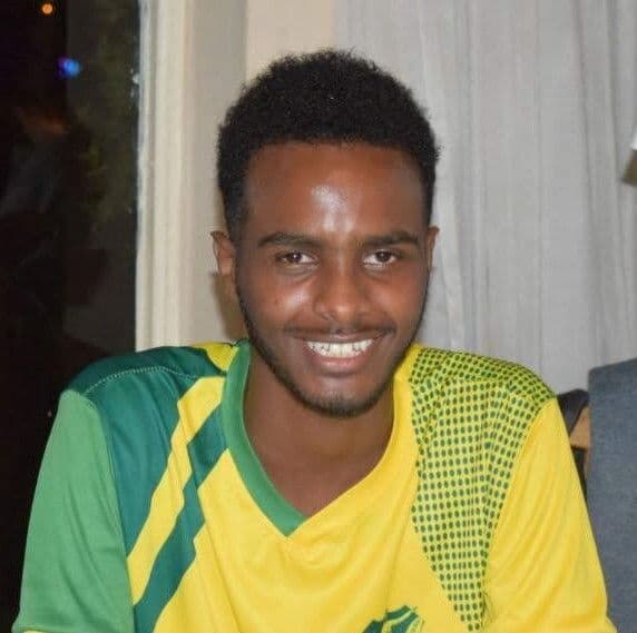 A headshot of Nuredin, a meidum/dark skinned Black man with short hair wearing a Brazillian soccer jersey and smiling into the camera. 