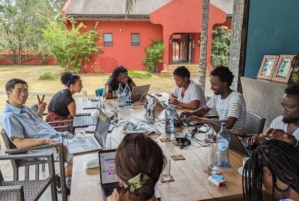 A photo of a subset of the DAIR team outside around a table, with many looking at their laptops. Nathan is looking into the camera and signing a peace sign with his hand.