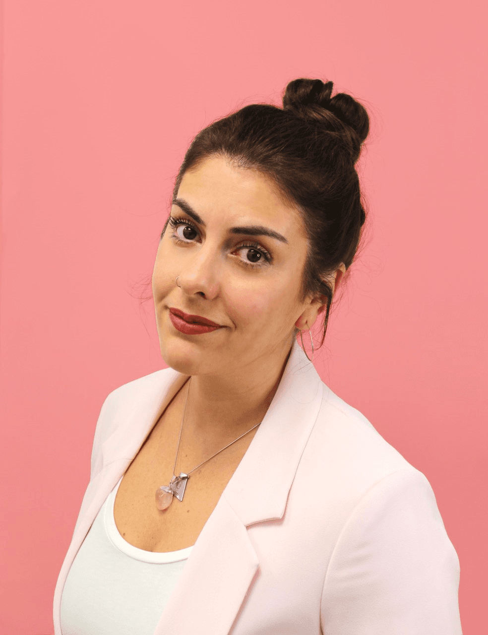 A picture of Mila looking into the camera, wearing a pink blazer and white shirt. And a necklace with a pink stone. Background is dark pinnk.