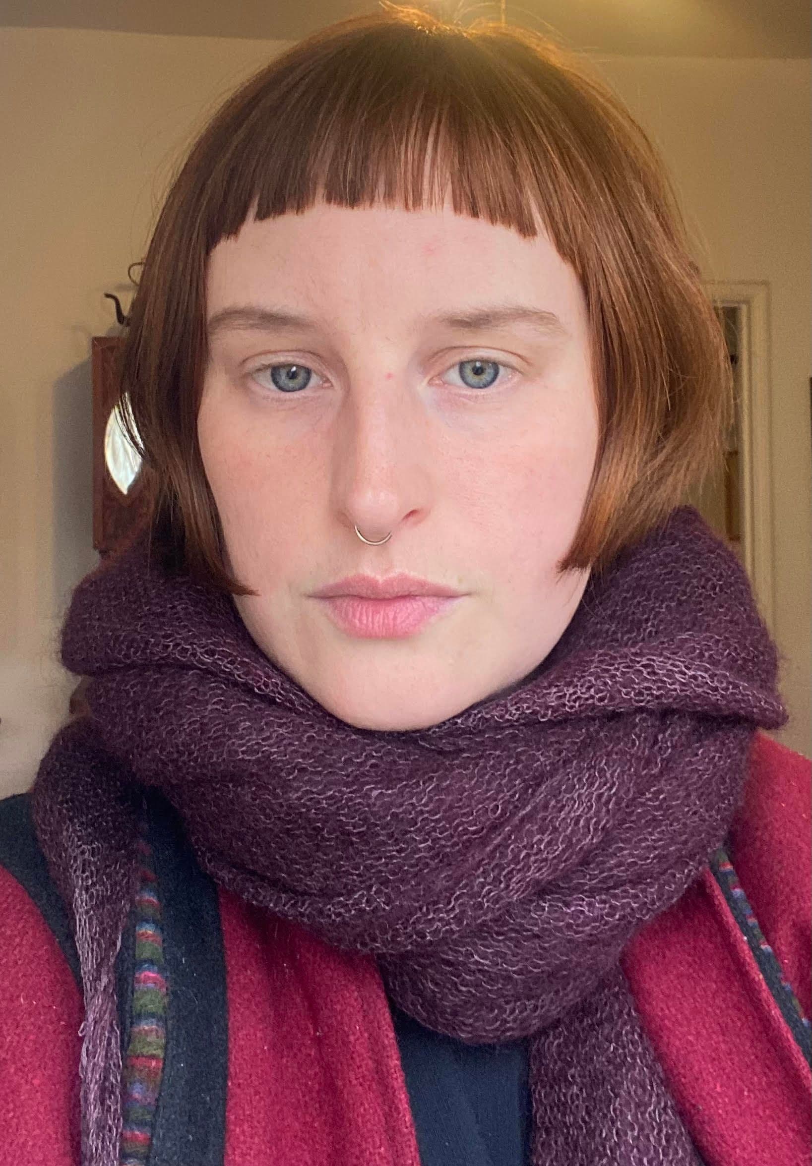 Headshot of a white person with straight red, chin-length hair and bangs. They are wearing a purple scarf and a pink shirt. The room behind her has off-white walls and is mostly obscured.