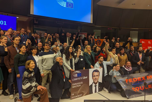 Photo of a crowd of 100 or so people lined up facing something off-camera on the right. Several have fists in the air, and those in the front row are holding signs. One sign shows a photo of Emmanuel Macron and says "Lobbyiste Uber: Emmanuel Macron" as if it were a business card.