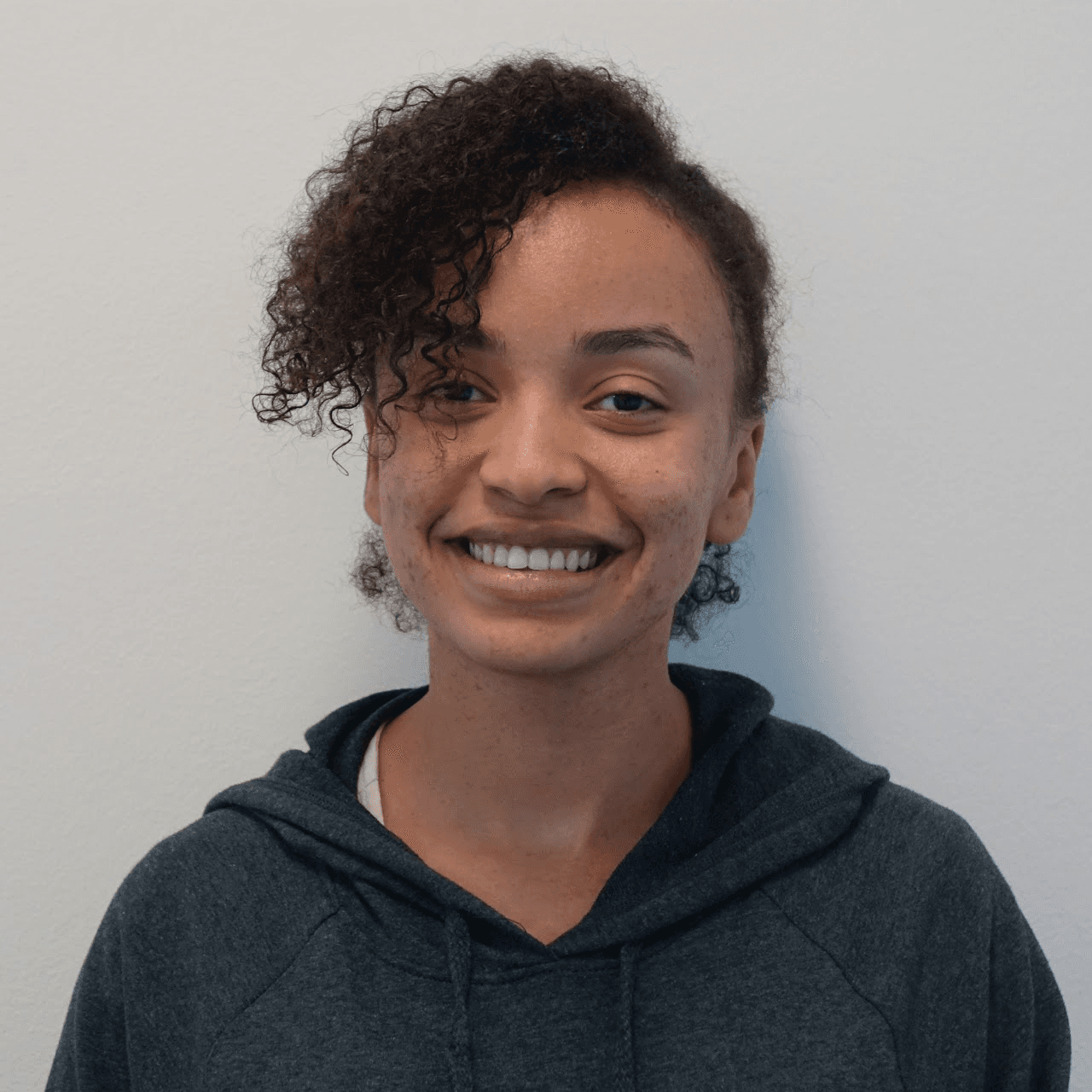 Photograph of a young Black woman from the shoulders up, with dark brown, curly hair sweeping over her forehead. She wears a gray hoodie. She is smiling, and standing with a white wall behind her.