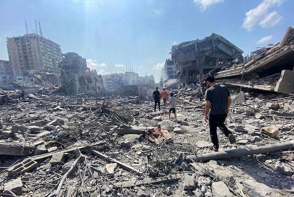 A photo of rubble in Gaza after an explosion, with 4 people walking through the rubble.