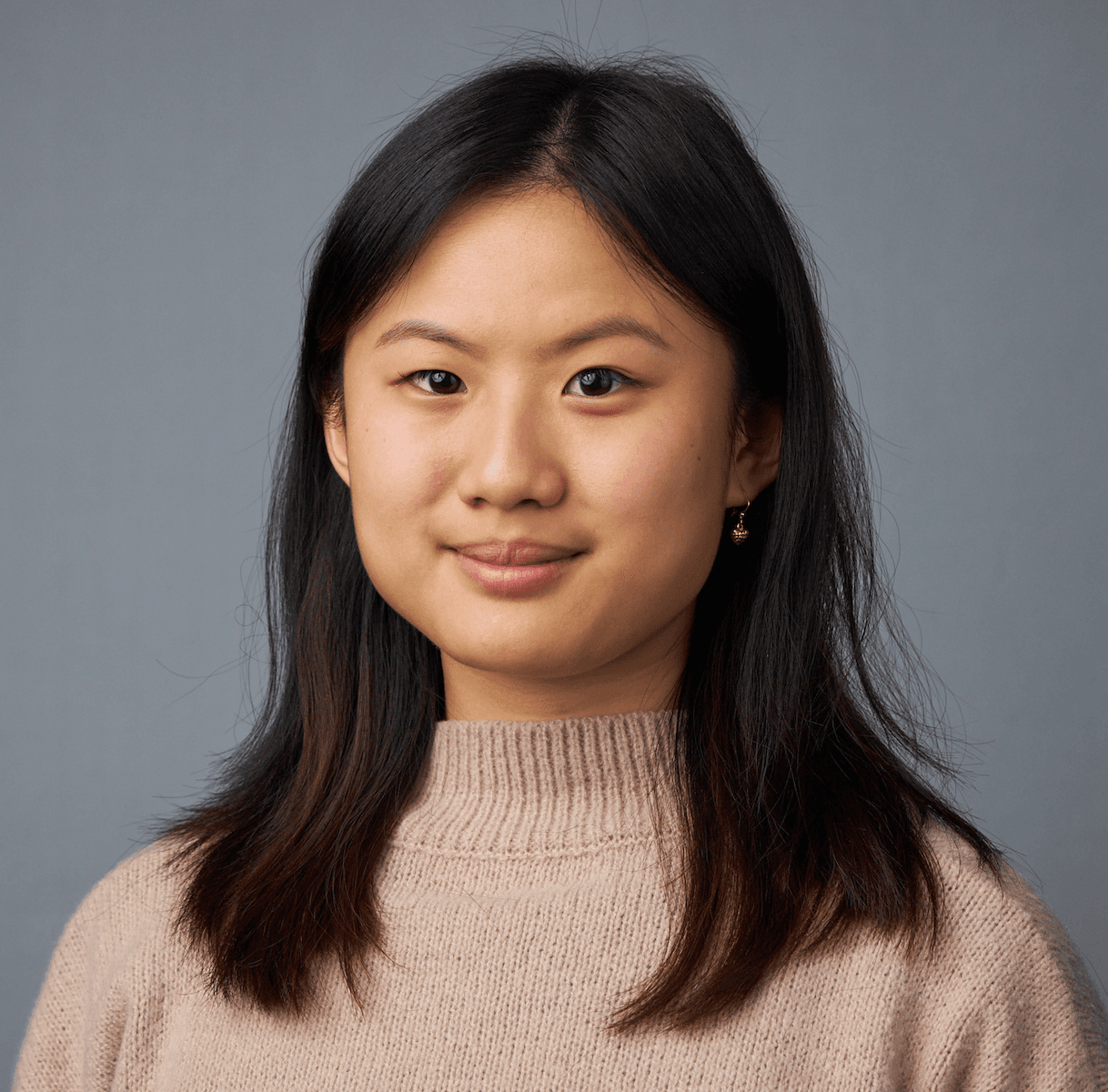 A headshot of an Asian woman with black hair past her shoulders. She is wearing a light brown sweater with a medium-high ribbed neck. She is wearing small earrings and smiling. In the background is a gray wall.