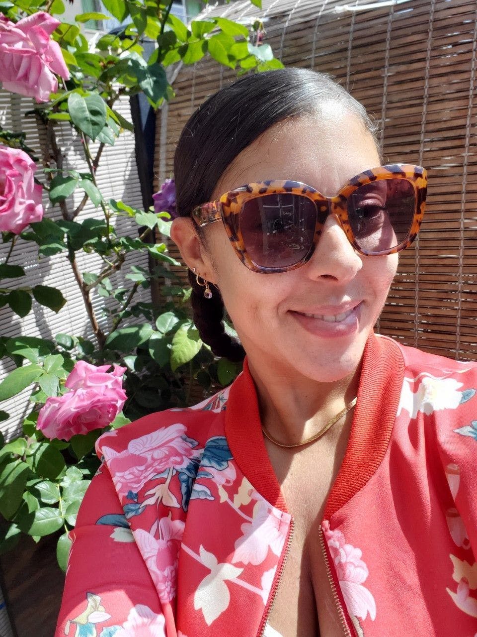 A headshot of Adrienne Williams, a light skinned Black woman with straight black hair that is pulled back. Wearing sunglasses, and a red jacket with colorful flowers, and pink roses in the background. 