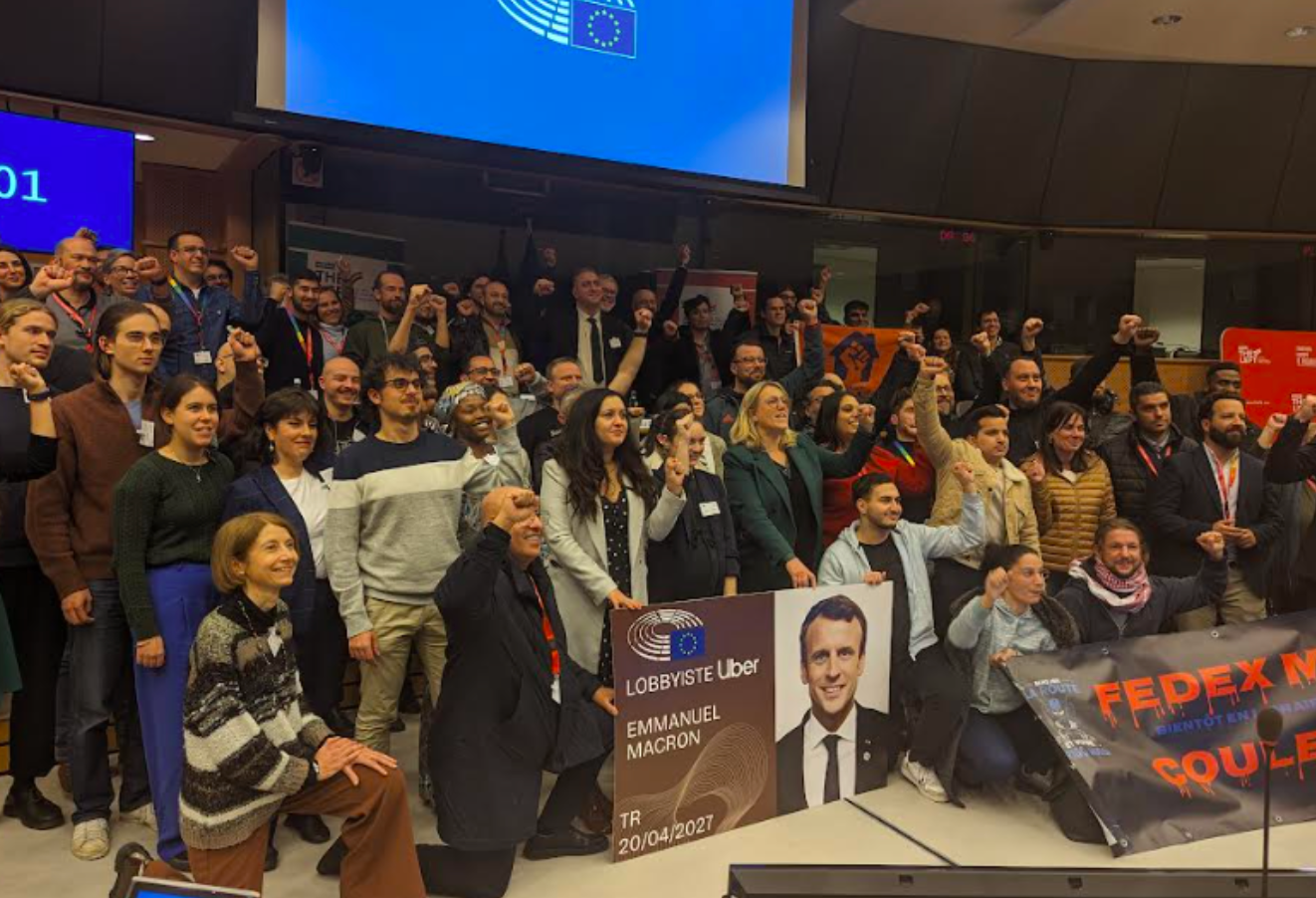 Photo of a crowd of several dozen activists in a large room. All are looking to the right, and several hold up fists. At the front are a couple signs. One reads, in French, "Lobbyiste Uber. Emmanuel Macron," and shows a photo of the French president. 