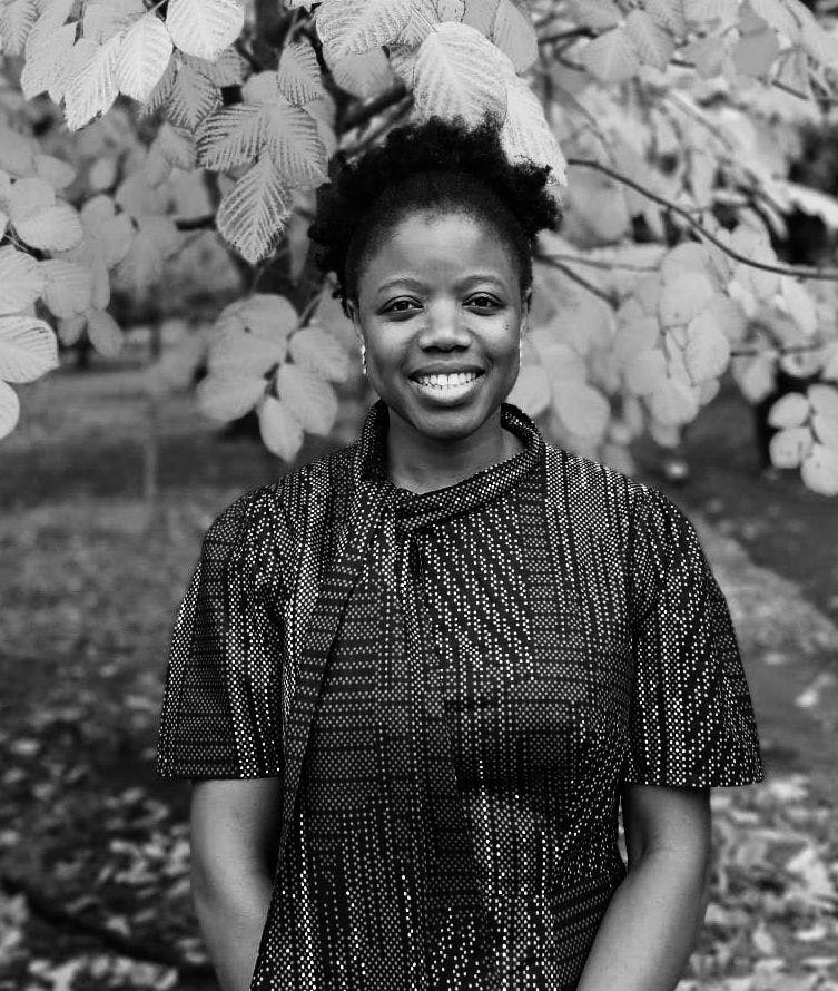 A black and white photo of a Black woman from the waist up. She has short black hair, and wears a short-sleeved garment with a variety of stripe patterns. She is standing outdoors, in front of a tree with pale leaves that show up as white.