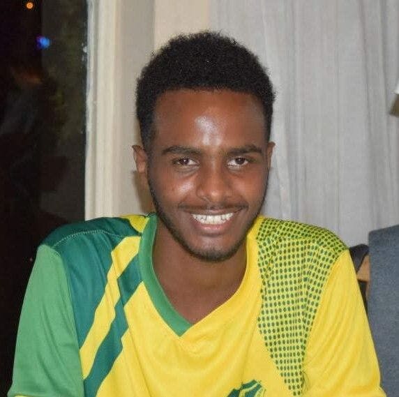 A headshot of Nuredin, a meidum/dark skinned Black man with short hair wearing a Brazillian soccer jersey and smiling into the camera. 