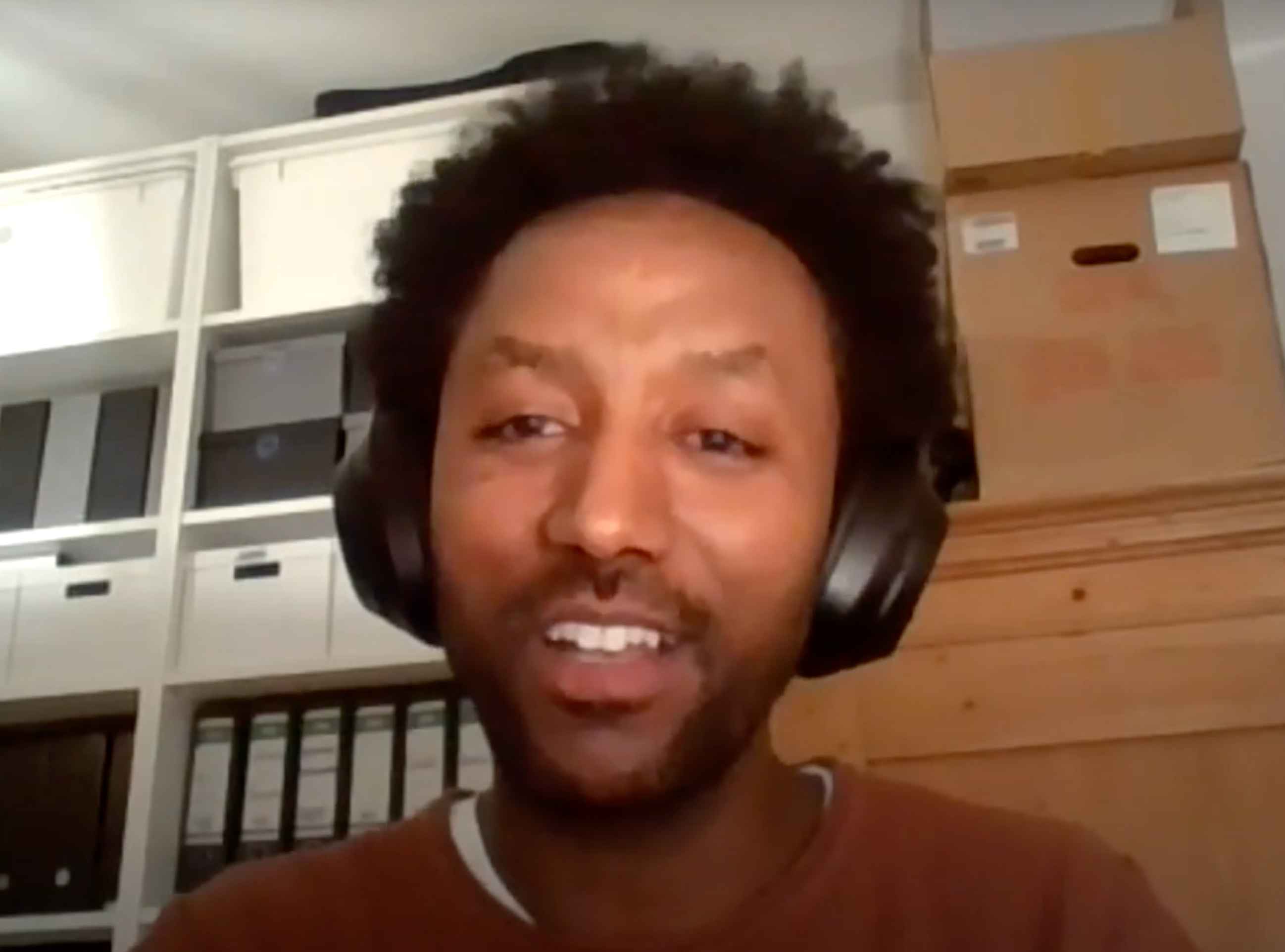 Asmelash Teka Hadgu, a Black man with medium, curly black hair, looks at the camera. He wears big, ear-covering headphones, and a reddish-brown shirt. Behind him are white storage shelves and a wooden cabinet stacked with boxes.
