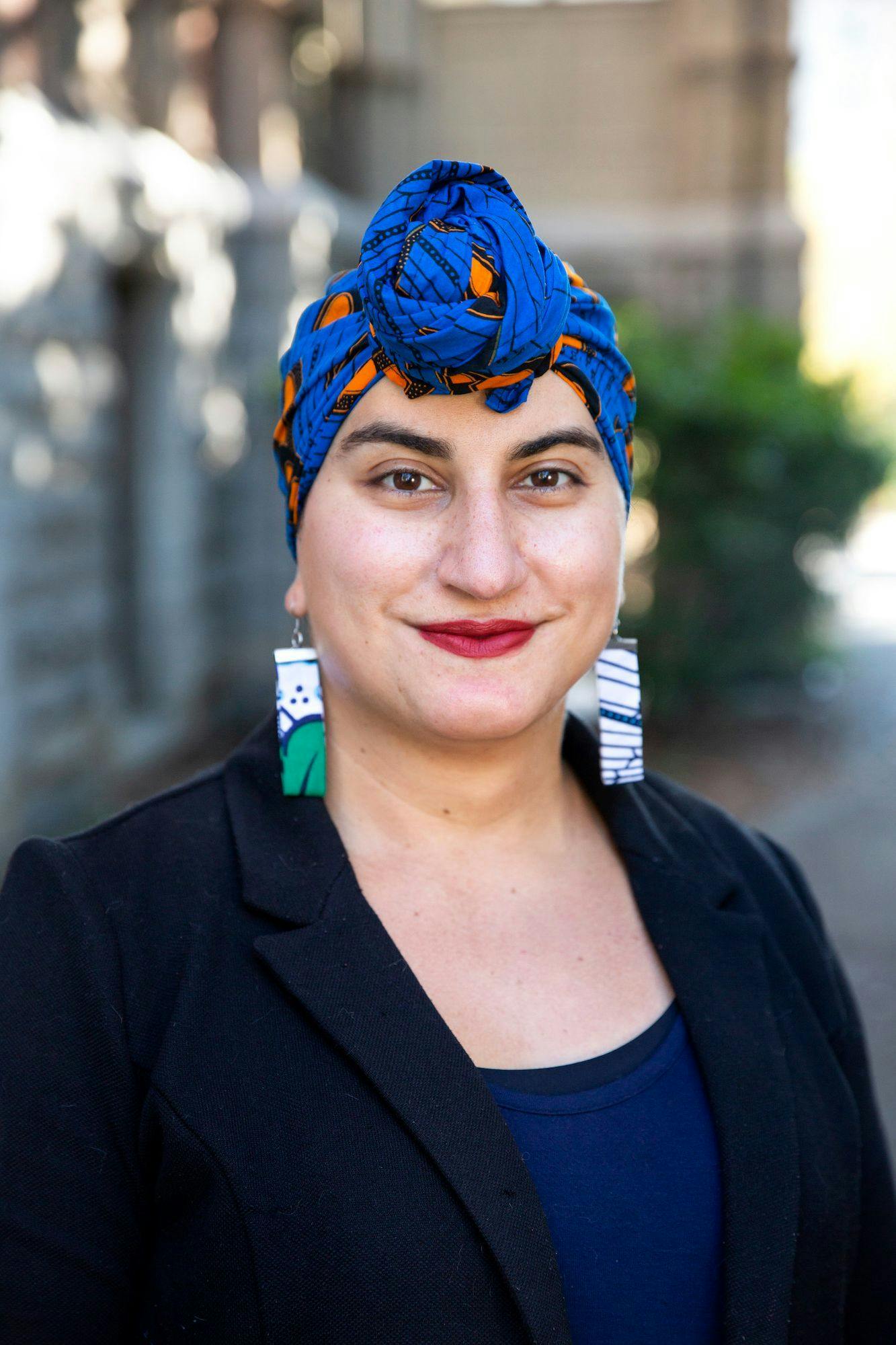 Headshot of Dr. Alex Hanna, an Arab woman, looking into the camera, wearing a blue headwrap, a pair of earrings and black blazer. 