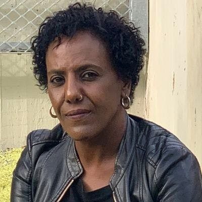 A headshot of Meron Estefanos, a Black woman with short hair, wearing small hoop earrings and a black top and leather jacket. In the background is a fence and grass which is slightly visible.  