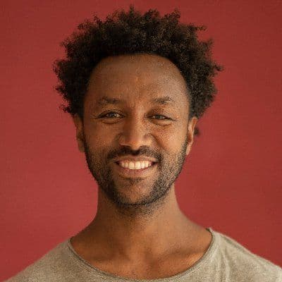 A headshot of Asmelash, a Black man with a short afro, wearing a cream shirt (background is red)