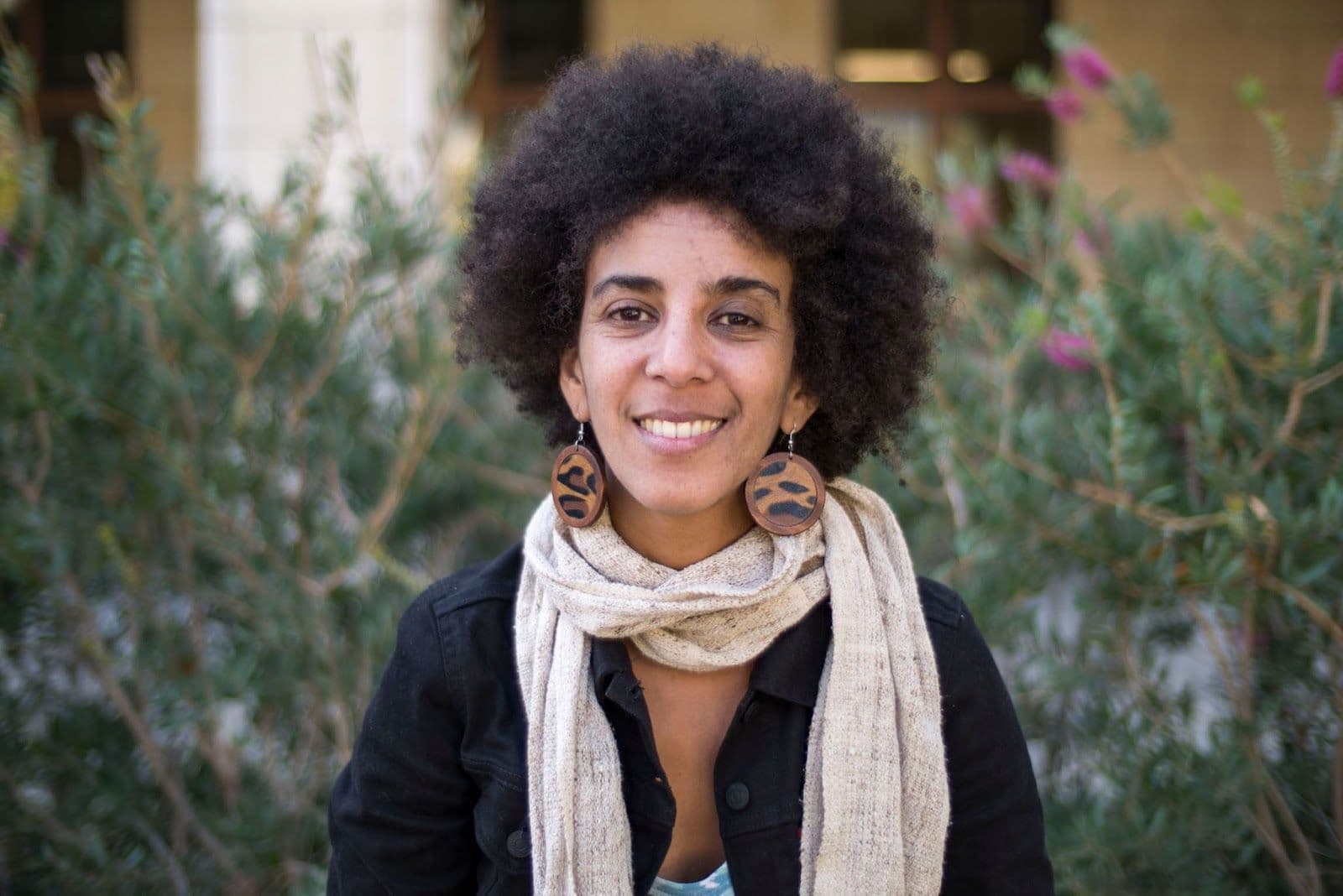 Timnit Gebru, a Black woman,  looking into the camera, wearing an afro, 2 large circular brown earings, a white scarf and a black sweater. Background is greanery. 