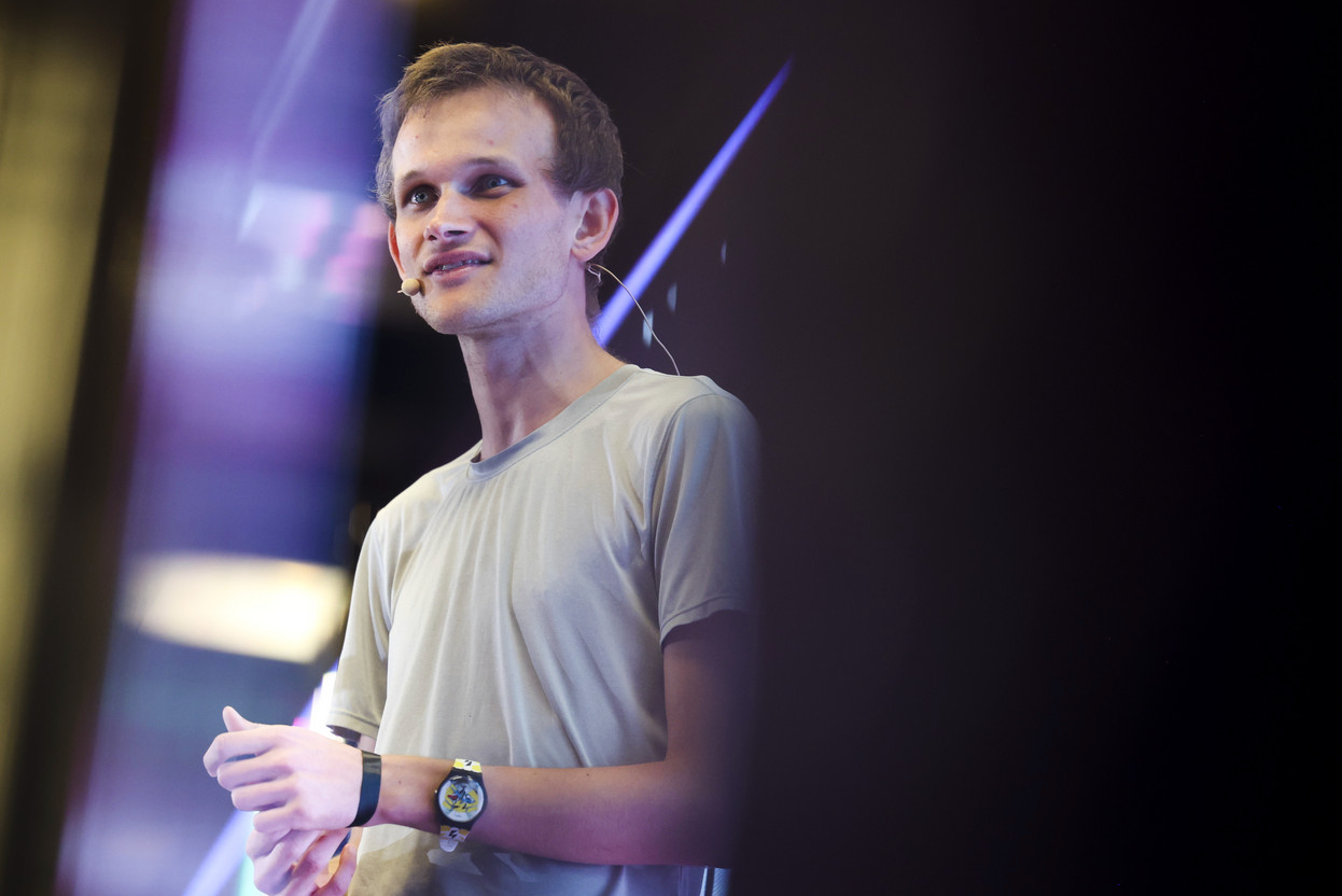 Waist-up photo of Vitalik Buterin, a white man with short brown hair. He is wearing an off-white t-shirt and a large watch on his left wrist. A beige-colored microphone rests on his right cheek next to his mouth. He appears to be speaking from a stage, but the background is out of focus: mostly black, with purple lighting.