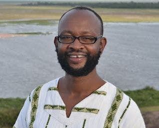 Dr. Ciira wa Maina, a Black man with short hair and a beard, wearing glasses, smiling into the camera. He's wearing a white shirt with green embroidery. Background is what seems like a lake and grass behind it. 