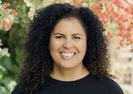 Dr. Safiya Noble, a Black woman, facing the camera smiling. She's wearing a black shirt, and her hair down. Orangish flowers and greenery in the background.